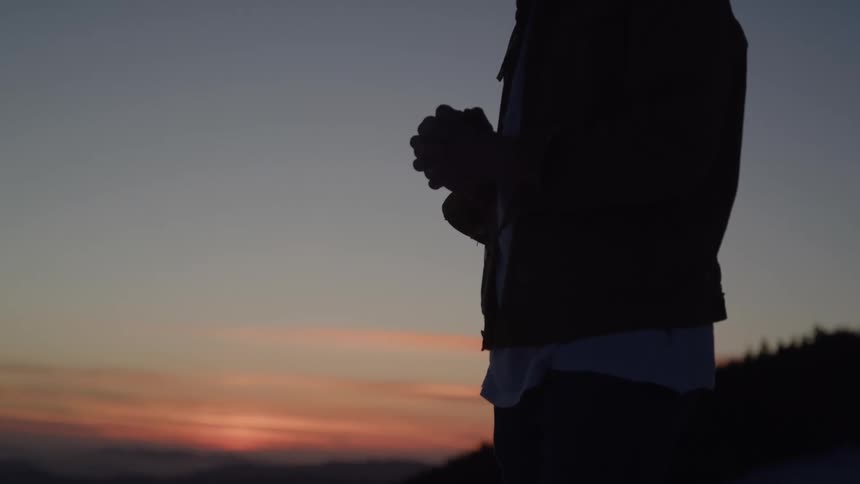 A close silhouette view shows a persons hands clasped together with a vibrant sunset and mountains visible in the background