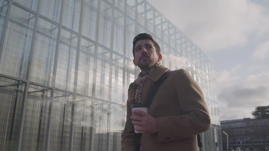 Man wearing brown coat and scarf holds coffee cup in front of glass building in urban setting