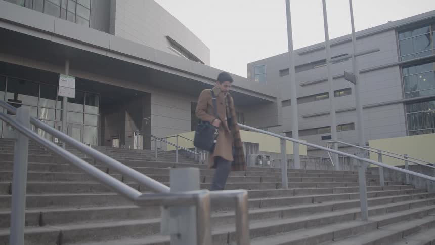 Person in a brown coat walks down concrete stairs in front of a modern building with large windows
