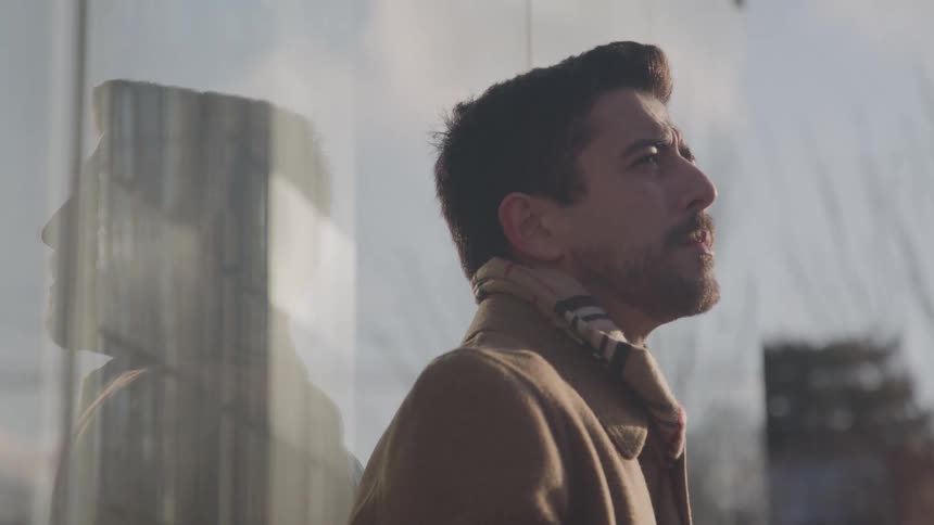 A side profile view shows a man with a beard wearing a camel coat and a patterned scarf standing outside a glass building and looking up with a thoughtful expression