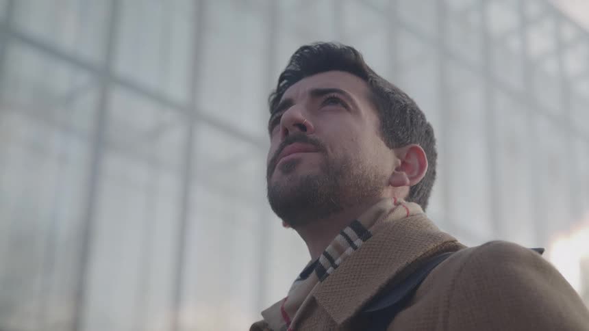A medium shot from a low angle captures a man with a beard and brown coat looking up and to the side he appears to be deep in thought or gazing at something in the distance