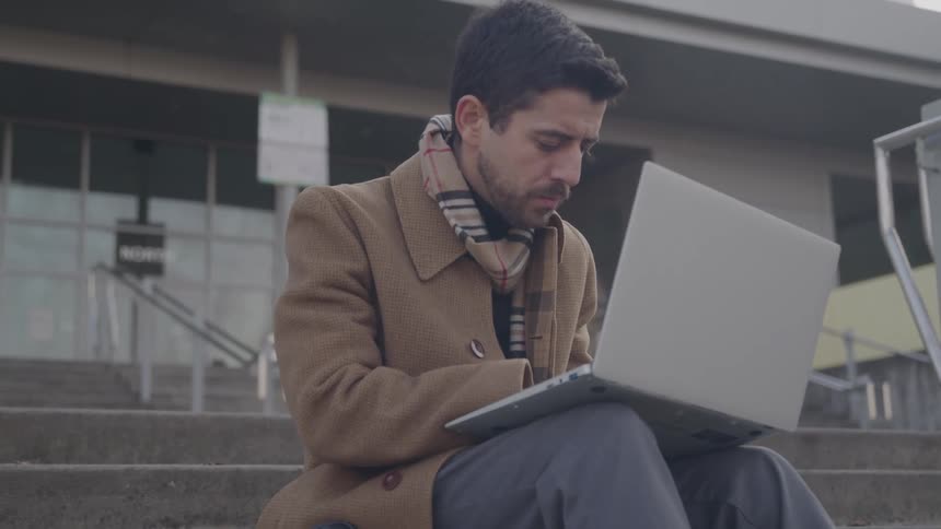Close up of a young man using a laptop while sitting outdoors on the stairs at the entrance of a building during daytime