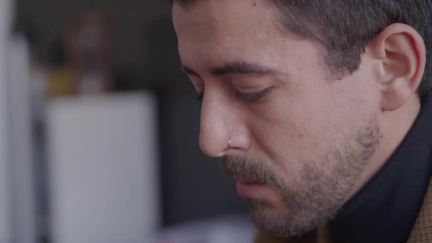 A young mans face is shown in a closeup from the side the man has a neutral expression and looking down in an indoor setting