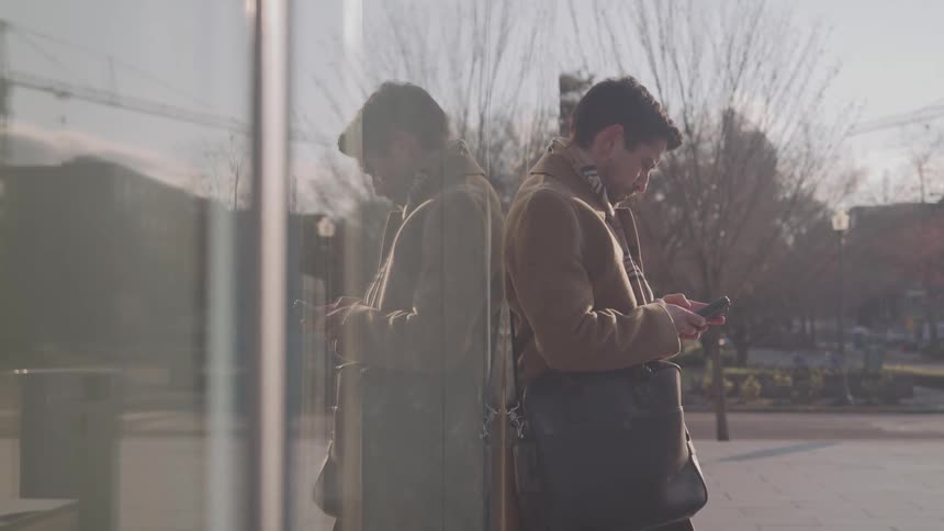 A close side view shows a man in a brown coat leaning against a glass building looking down at his phone with a black satchel over his shoulder