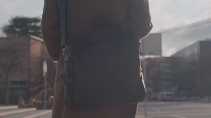 A camera follows a man wearing a long camel colored coat and boots as he walks away on a sunlit sidewalk casting a long shadow