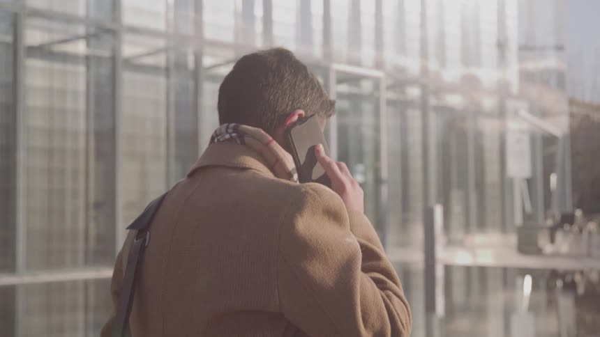 A man wearing a camel coat and a patterned scarf is seen from behind walking and talking on a smartphone outside a modern glass office building on a sunny day