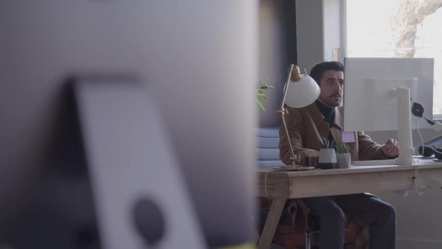 A young man working with a desktop computer while sitting at a desk in an office during daytime