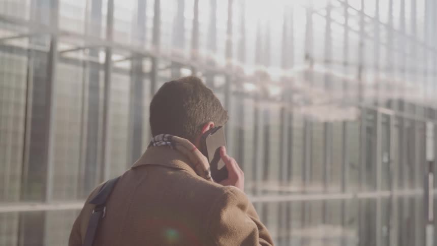 A young woman wearing a brown coat holding a phone to her ear walking along the sidewalk beside a towering office highrise under the bright midday sun