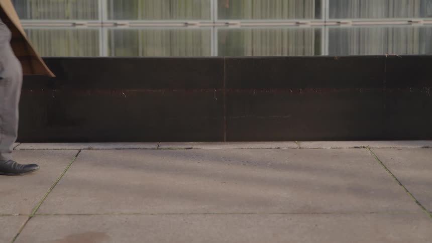 Person in gray pants and black shoes walks briskly on sidewalk in front of glasspaneled building