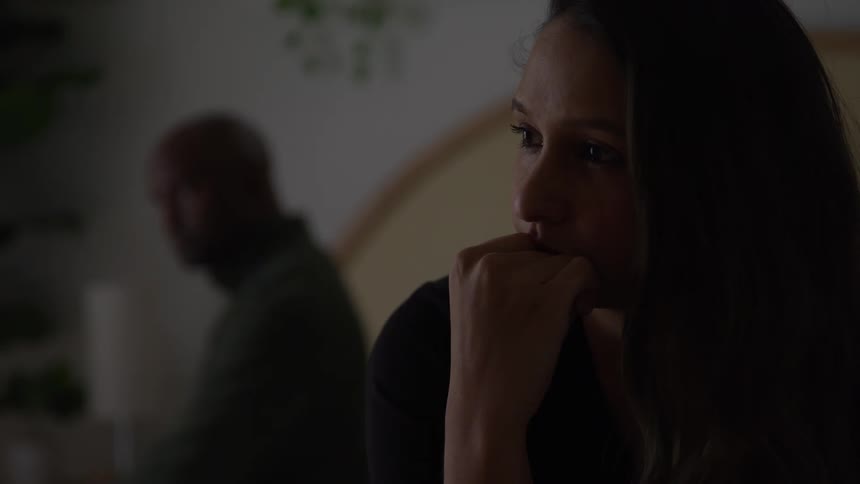 The face of a woman with long hair resting her hand on her chin with a man out of focus in the background sitting in a room with low light