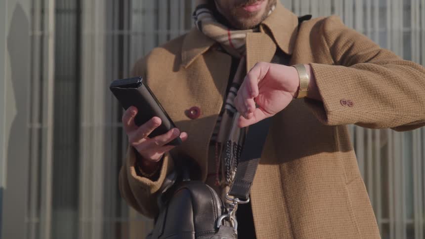 Man in a coat and scarf checks the time while holding a phone and bag