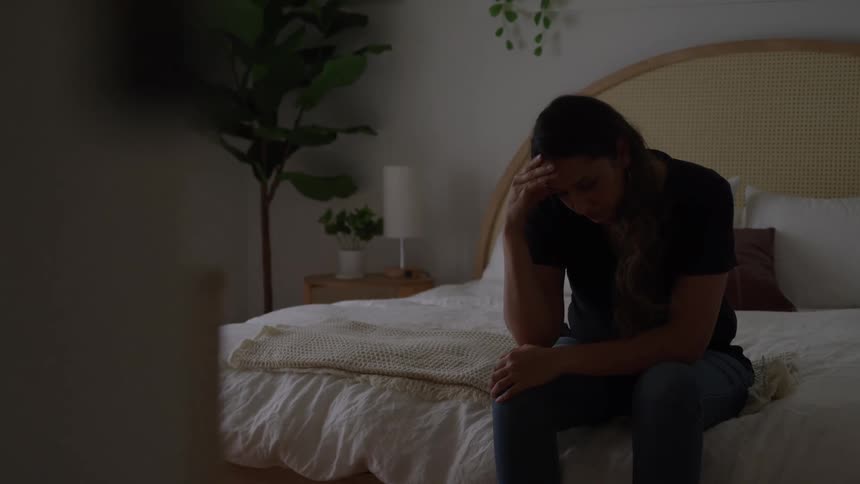 Person seated at the edge of the bed remains leaning forward with hand on forehead in lowlight setting