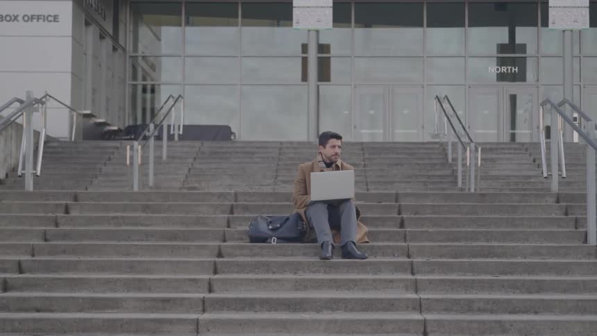A man with a beard wearing a brown coat and jeans is sitting on a large outdoor concrete staircase working on a laptop with a bag next to him