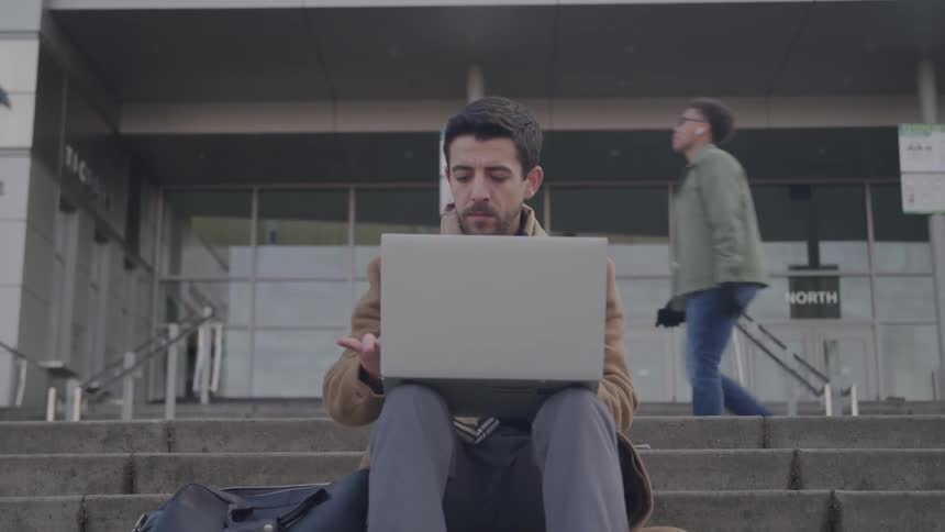 Close up of a young man using a laptop computer while sitting outdoors on the stairs at the entrance of a building during daytime