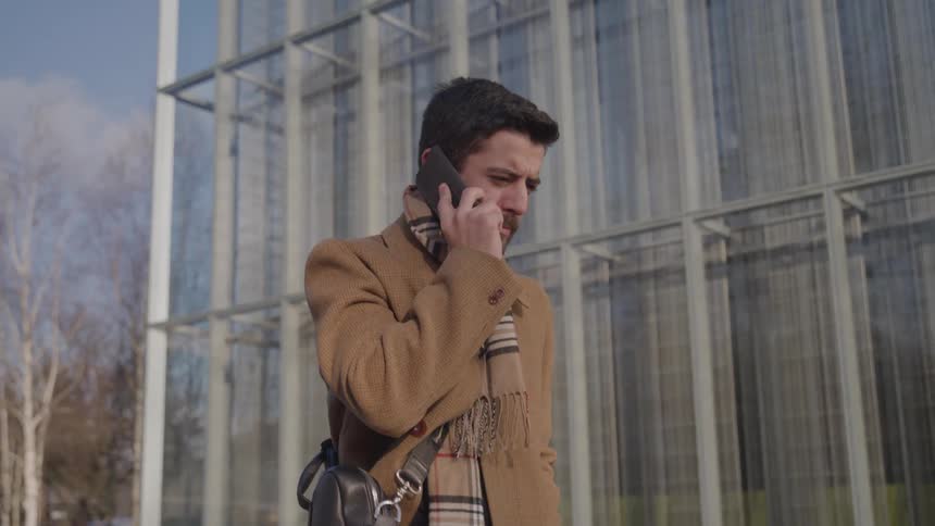 The man talks on his phone will walking past the glass windows of the building