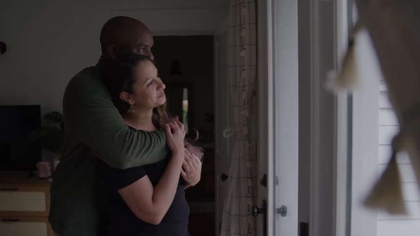 A man puts his arm around a woman and hugs her from behind as she looks out of a glass door