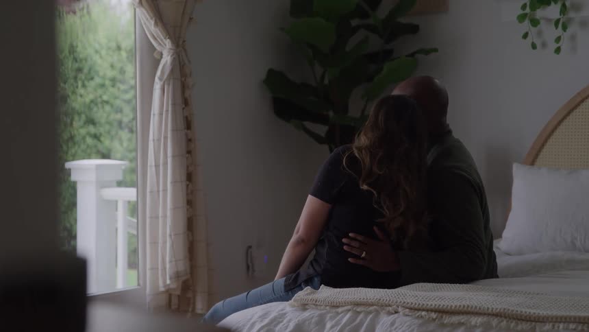 A loving couple sits on their bed by a window gazing outside while embracing each other