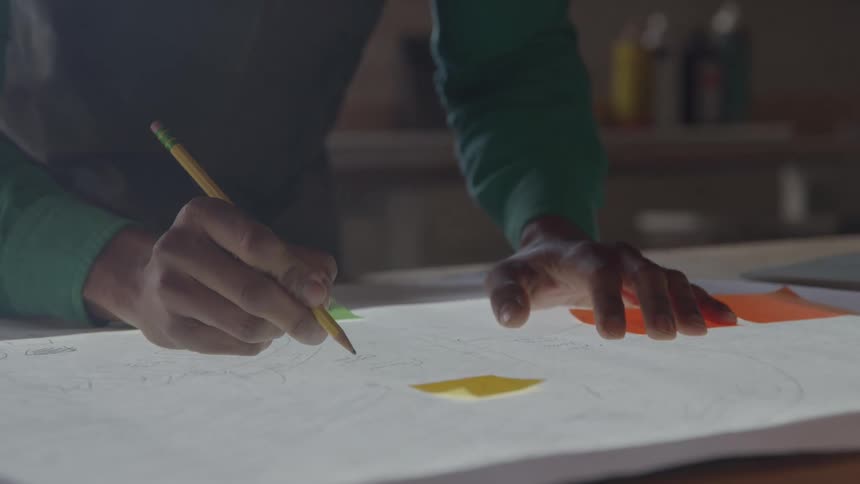 A close view shows a persons hands wearing a green shirt using a pencil to trace a design on a glowing paper on a light table with small colorful sticky notes nearby
