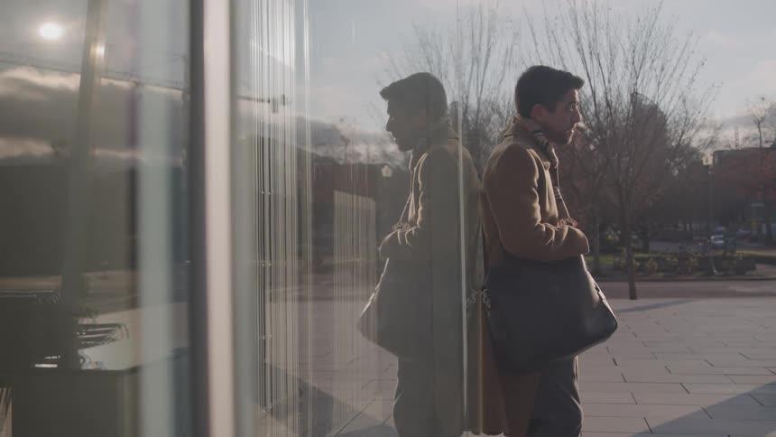 A young man standing outdoors in front of a glass building in a suburban city during daytime