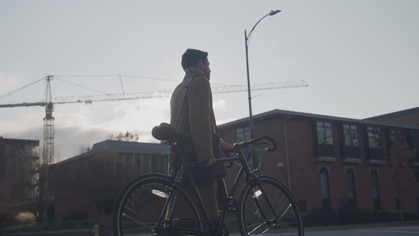 A man stands on a crosswalk holding a bicycle while sunlight shines behind him with buildings and a crane in the background