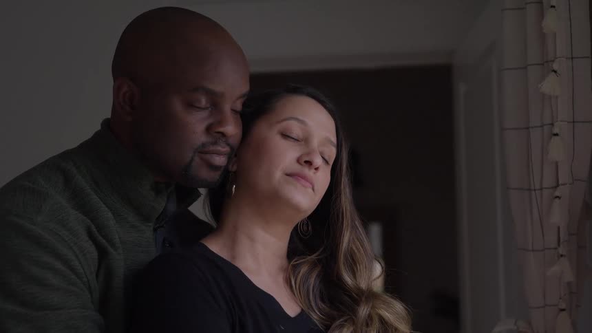 Close up portrait of a black man romantically hugging his female partner from behind as they stand in a room during daytime