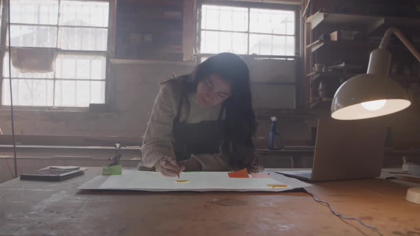 A young woman diligently works on a design project at a large wooden desk sketching and using a laptop in a sunlit workshop