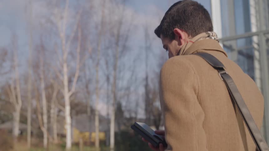 A young man is walking on a sidewalk while swipping on his smartphone wearing a sweater and with a bag