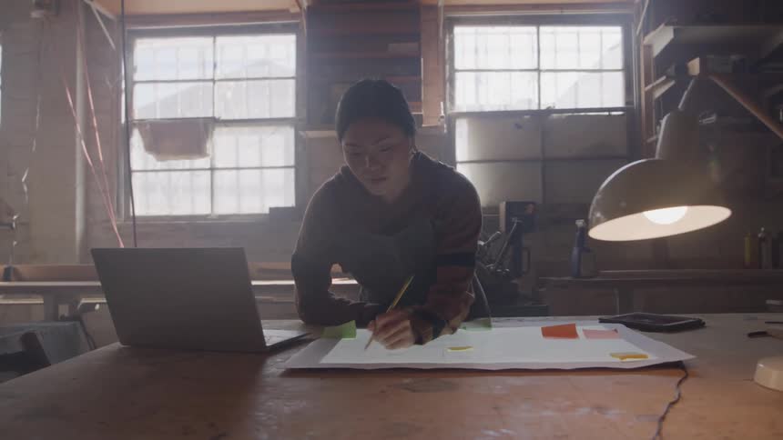 A young woman is working at a desk inside a warehouse at midday writing with a pen on a todo list while a laptop sits beside her