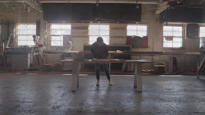 Person in a workshop lit by windows leaning over a large wooden table in the center of the space