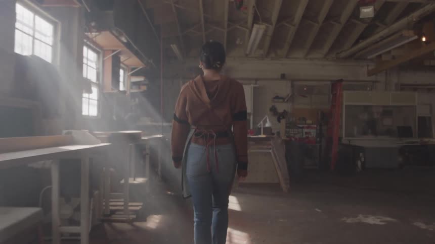 A woman with a short ponytail wearing a brown shirt and blue jeans walks into a dusty sunlit woodworking workshop with tools and workbenches visible in the background