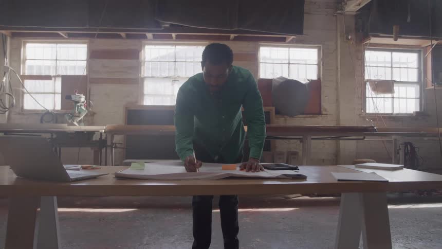 Man in a workshop drawing designs on a large table with a laptop and papers around