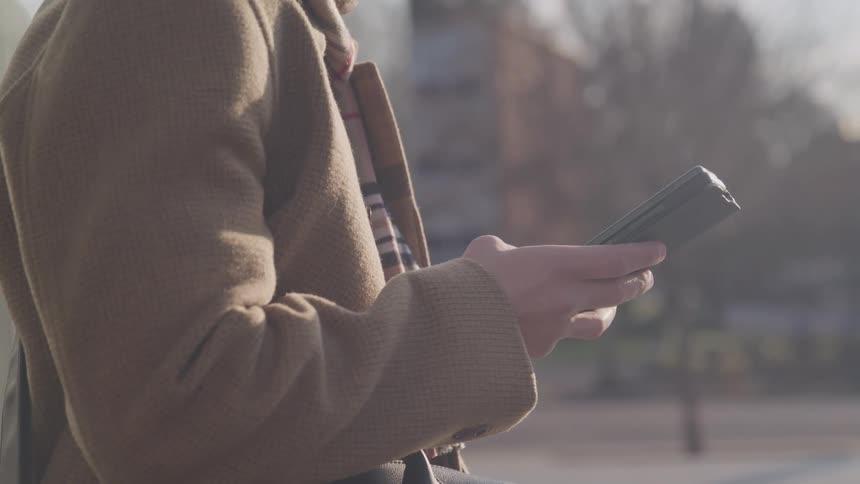 A person in a brown coat and plaid scarf checks their smartphone outdoors
