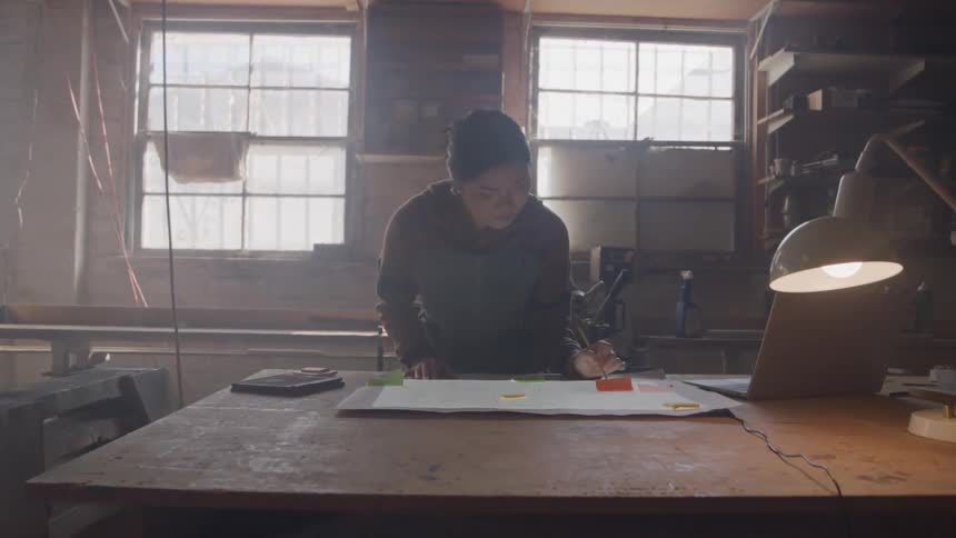 Woman works focused at wooden table with drawn plans laptop and office supplies in space lit by natural light
