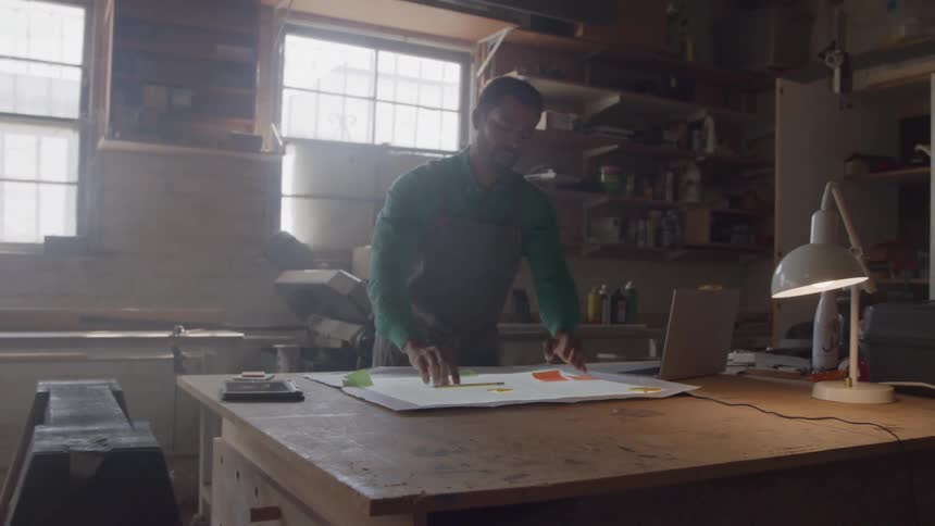 A man in a workshop stands over a lighted work table drawing with a pencil on a large sheet of paper with a laptop and a lamp visible on the table