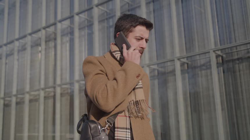 A man in a camel coat and scarf holds a phone to his ear with a furrowed brow looking focused