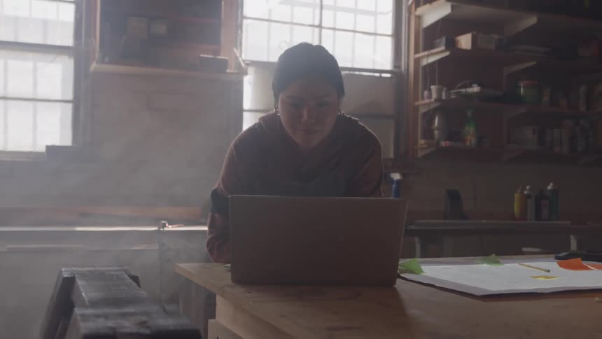 Woman works on a laptop inside a workshop lit by the window