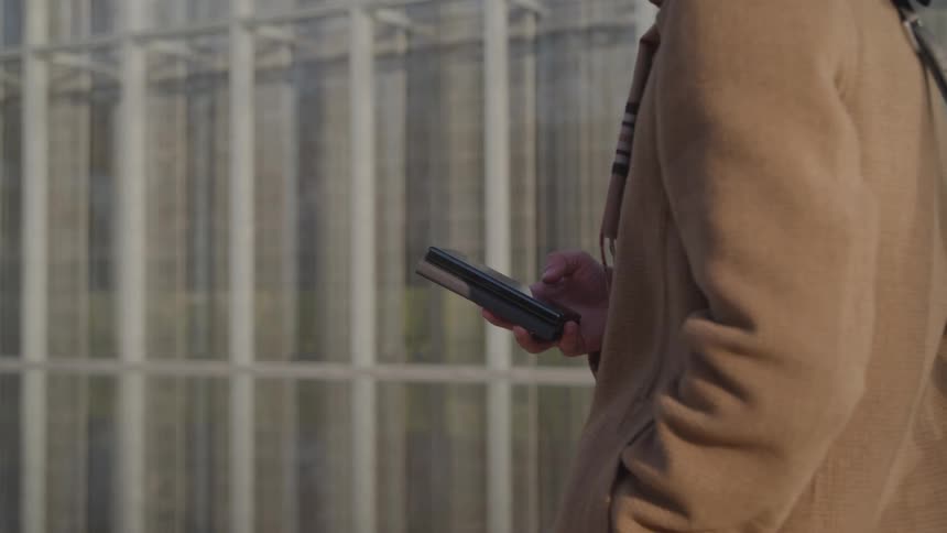 A close side view shows a man in a brown coat with a bag over his shoulder walking and scrolling on a smartphone