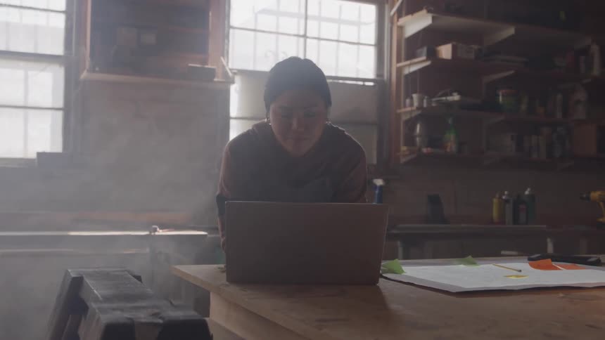A person sits at a workbench and looks down at a laptop screen
