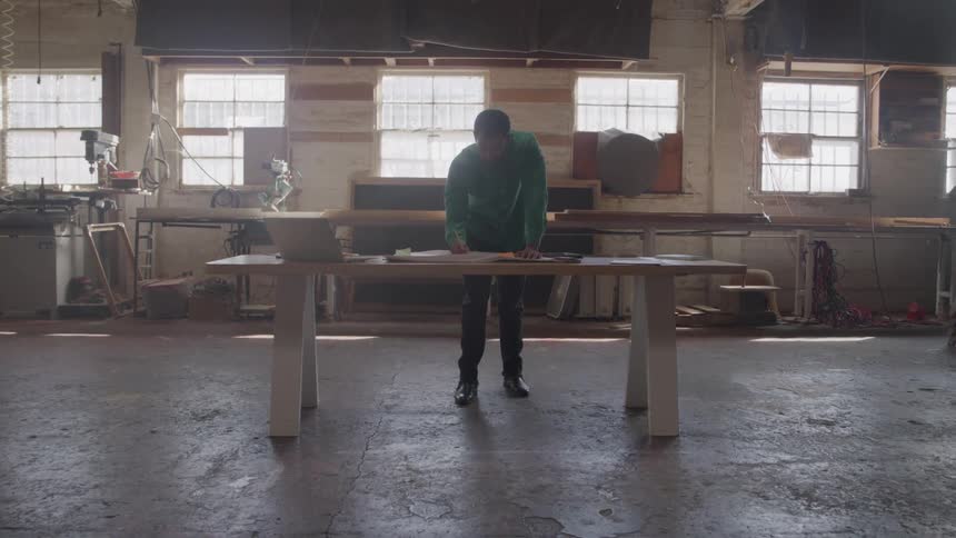 A person stands and leans over a workbench in a workspace with multiple windows