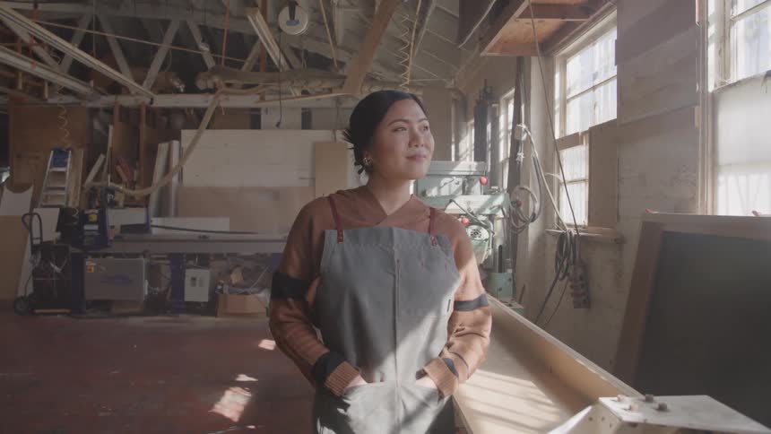 A woman stands with her hands in her pockets in a woodworking workshop