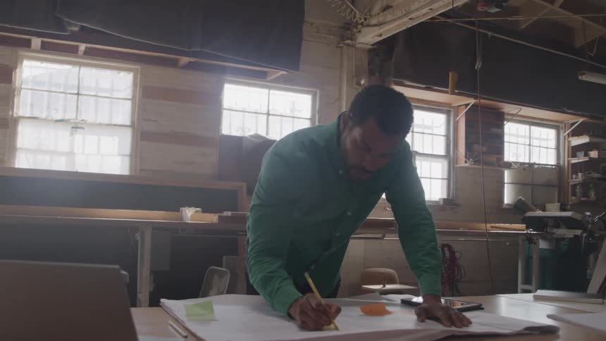 The man walks over to the blueprints and picks up a pencil