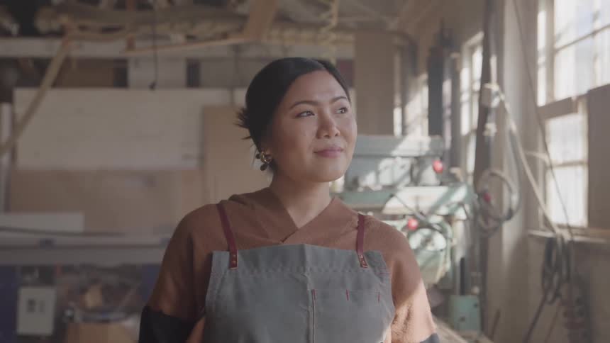 A proud female artisan in a workshop smiles and looks off to the side surrounded by tools and equipment