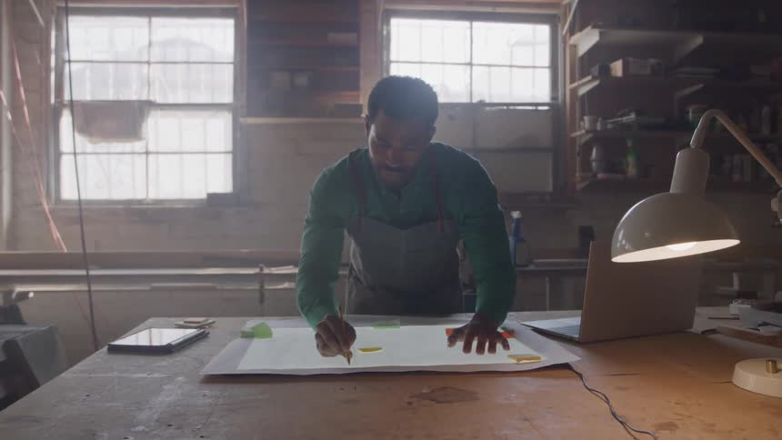 Man working on a project at the table drawing on a large sheet of paper with a laptop and lit lamp beside him