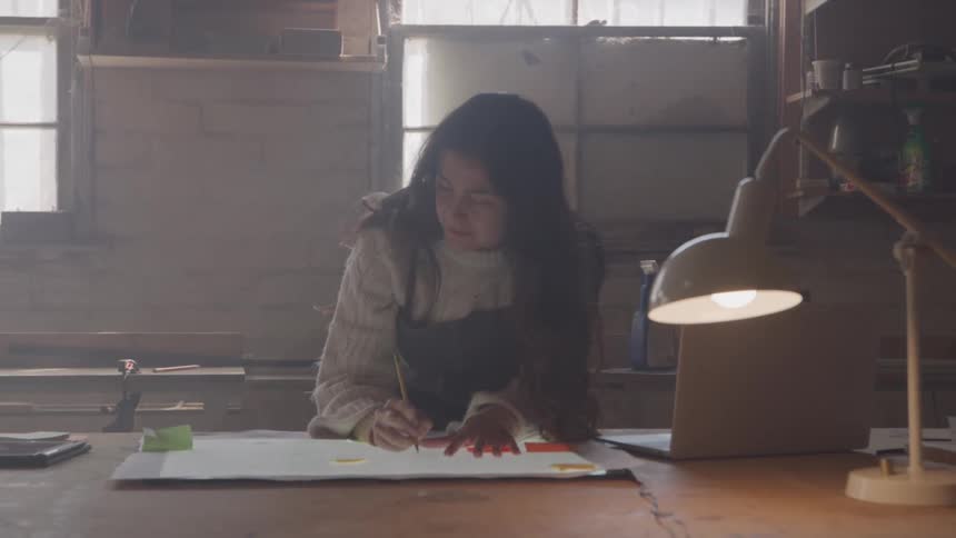 A woman sits at a desk in a workshop and draws on a blueprint