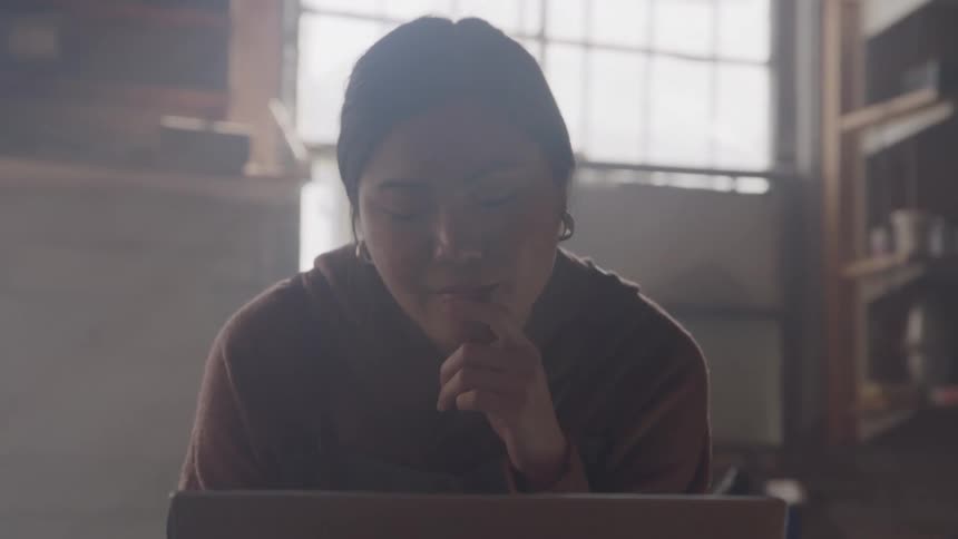 A young woman sits at a desk looking at a laptop screen with one hand resting on their chin