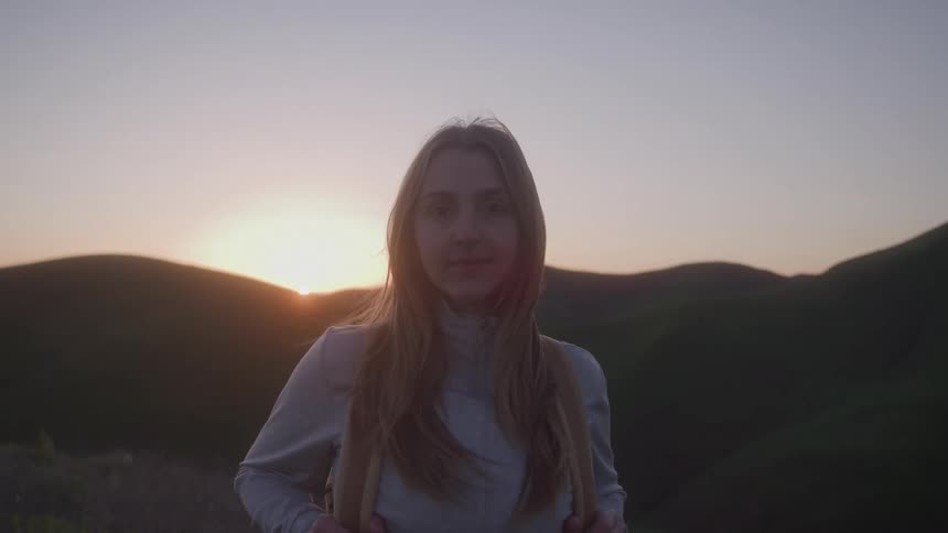 Blonde woman with a backpack looks at the horizon at sunset in the mountains