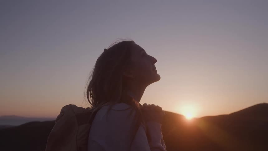 Woman with backpack gazes at the horizon at dusk in mountainous landscape lit by golden sunset light