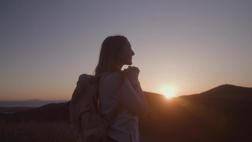 A woman stands on a hill looking at a sunset with mountains on the horizon