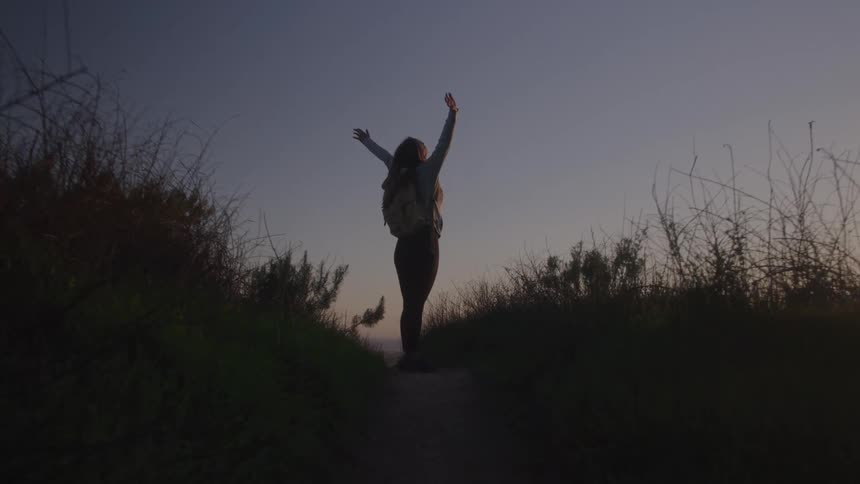 Person with a backpack walking on a trail at sunset with arms raised