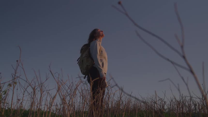 A woman with a backpack and a hoodie stands on a hill looking up at the sky while surrounded by grass and weeds